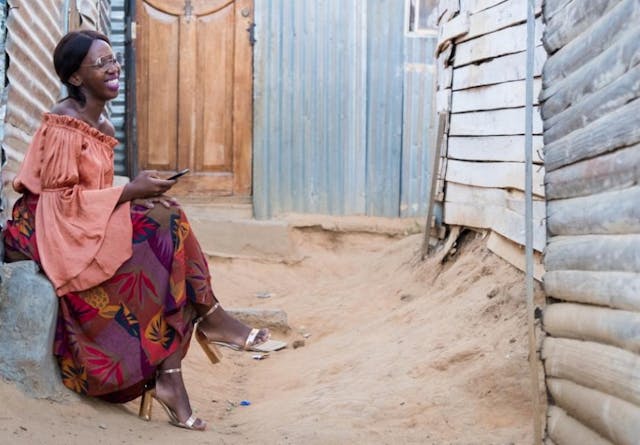 A well-dressed black woman with eye-glasses sits by her shack and uses her mobile phone