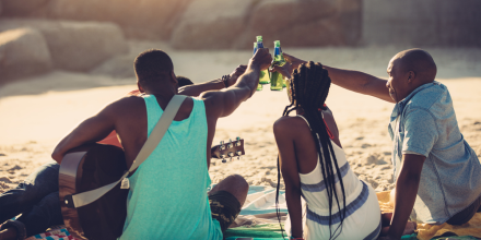 Young people toasting with beer bottles while sitting on beach
