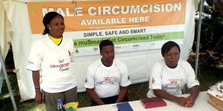 Three community workers sit at a table with a sign behind them that reads 'Male circumcision available here'