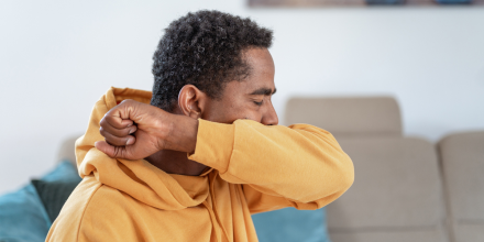 Adult man wearing a yellow hoodie in a living room, coughing into elbow