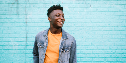 A smiling African American man stands in front of a blue wall wearing nice casual clothing.