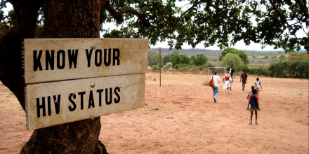 Shot of rural area with a few people walking on it and a sign on a tree says 'Know your HIV status'