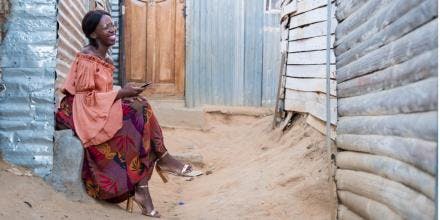 A well-dressed black woman with eye-glasses sits by her shack and uses her mobile phone