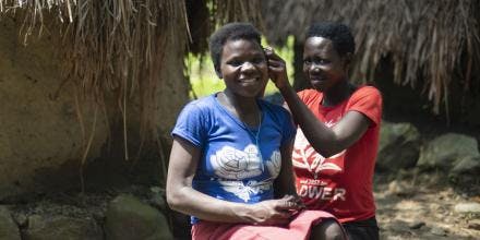 A Ugandan woman helps another woman with her hair while sitting outside