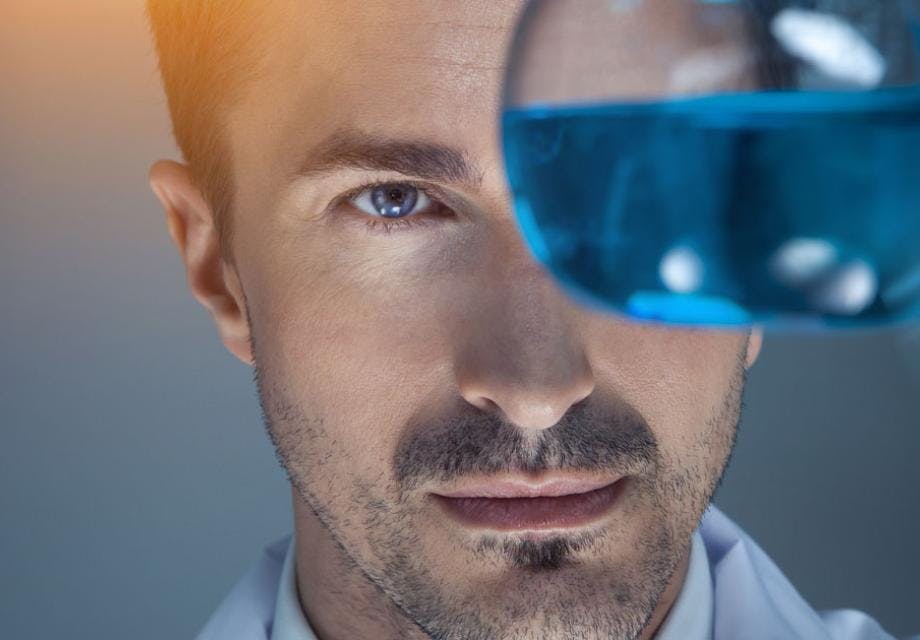 A chemist holding up and looking at a glass beaker with blue liquid inside