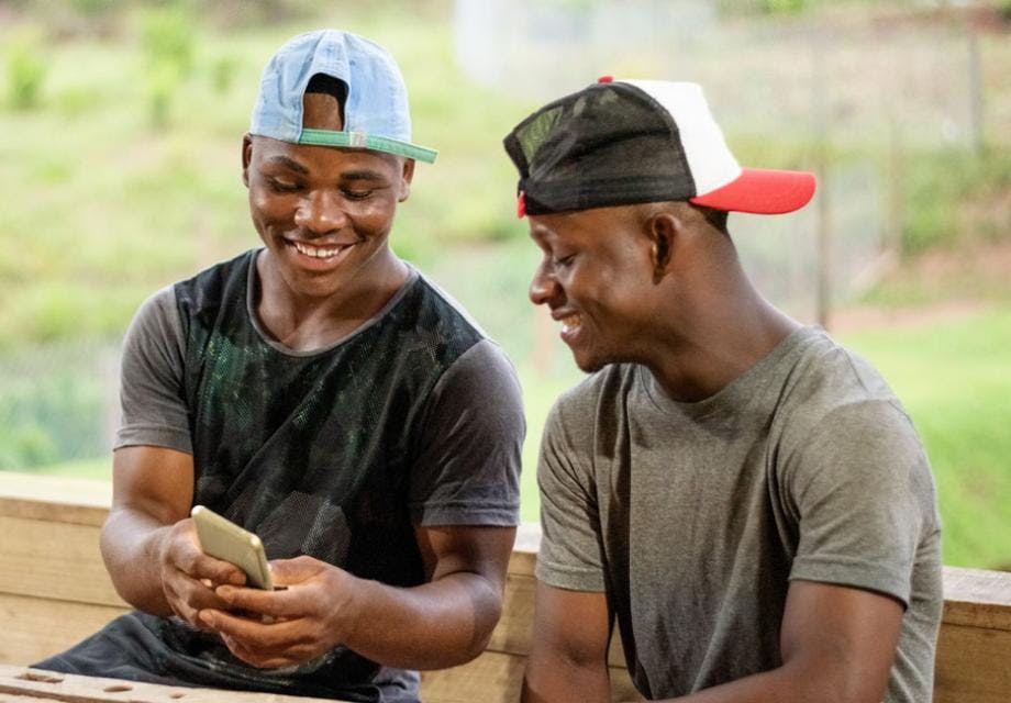 Two young men sit at a table while looking at a mobile phone and smiling