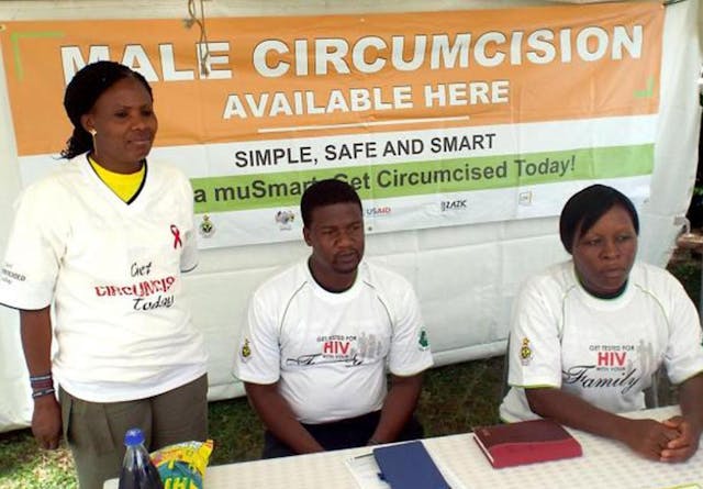 Three community workers sit at a table with a sign behind them that reads 'Male circumcision available here'