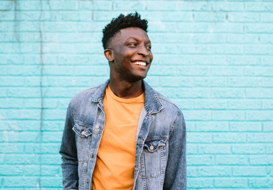 A smiling African American man stands in front of a blue wall wearing nice casual clothing.