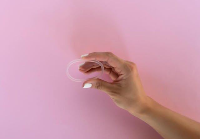 A woman's hand holds a vaginal ring in front of a pink background