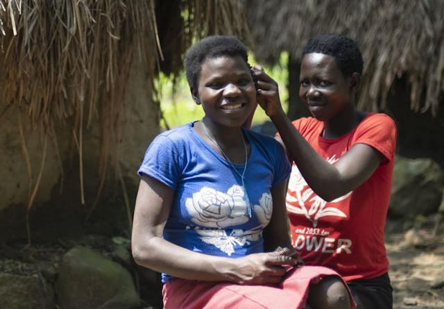 A Ugandan woman helps another woman with her hair while sitting outside