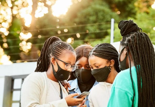 Teenage friends with face masks looking at a smartphone together outdoors